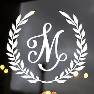 May include: White vinyl decal of a stylized letter M surrounded by a wreath of leaves. The decal is on a dark background.