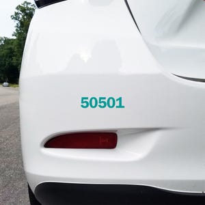 May include: A close-up of the rear of a white car with the teal number "50501" displayed on the bumper. The car's taillight is visible, and the background shows a blurred outdoor scene.
