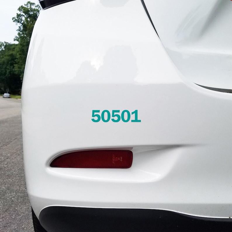 May include: A close-up of the rear of a white car with the teal number "50501" displayed on the bumper. The car's taillight is visible, and the background shows a blurred outdoor scene.