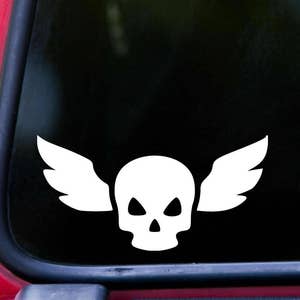 May include: White vinyl decal of a skull with wings. The skull has two eye sockets and a nose. The wings are spread out on either side of the skull.