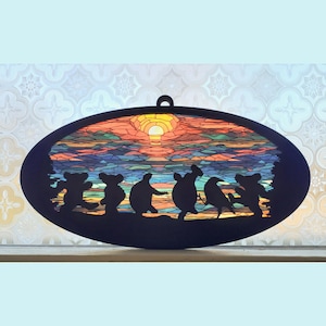 May include: A stained glass oval wall hanging with a sunset scene and a silhouette of seven bears dancing. The stained glass features a variety of colors, including red, orange, yellow, blue, green, and purple.