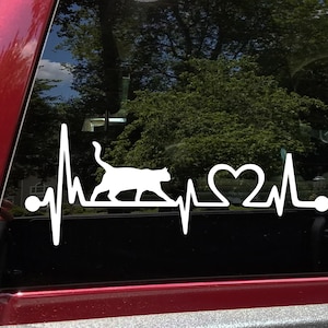 May include: White vinyl decal of a cat walking with a heart and a heartbeat line. The decal is on a black car window.