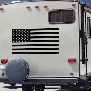 May include: A black and white American flag decal is affixed to the back of a recreational vehicle. The flag features 50 white stars in a blue rectangle and 13 alternating black and white stripes. The RV has a spare tire cover and red tail lights.