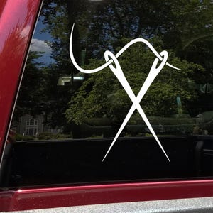 May include: A white vinyl decal of two crossed needles with thread, and a curved needle with thread. The decal is applied to a red vehicle window. The background shows trees and a house.