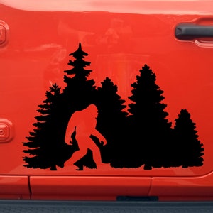 May include: A black vinyl decal of a Bigfoot walking through a forest of pine trees. The decal is applied to the side of a red Jeep Wrangler.