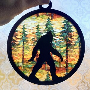 May include: A round stained glass window with a black frame depicting a silhouette of Bigfoot walking through a forest. The forest is depicted with a colorful stained glass design of trees and foliage.