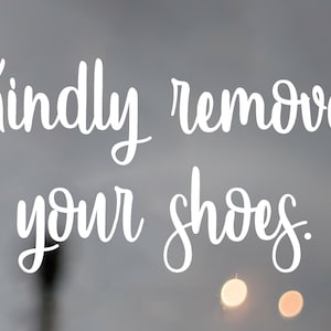 May include: White cursive text on a gray background reads "Kindly remove your shoes." The sign is displayed on a glass surface, with a wooden frame on the right side. Soft, blurred lights are visible.