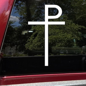 May include: A white Chi Rho symbol, a Christian monogram, is displayed on a vehicle window. The symbol, a combination of the Greek letters Chi (X) and Rho (P), is a prominent religious emblem. The background shows a view of trees and a house.