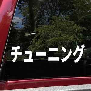 May include: White Japanese text on a car window that reads "チューニング" (Chūningu), meaning "tuning". The background shows a blurred view of trees and a red car frame.
