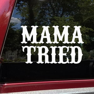 May include: White vinyl decal with the words "MAMA TRIED" in a decorative font. The decal is affixed to a window, with a blurred outdoor scene visible through the glass. The decal is likely for a car or truck.