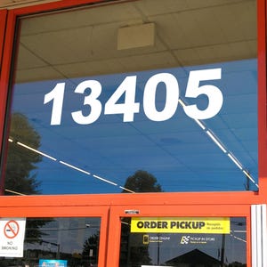 May include: A red storefront window with the number 13405 displayed in white. The window reflects a blue sky and trees. A sign below the window reads "ORDER PICKUP" with the text "Recogido de pedidos" in smaller font. The sign also includes the options "ORDER ONLINE" and "PICKUP IN STORE".