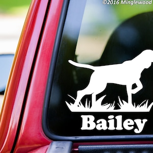 May include: White vinyl decal of a dog silhouette walking through grass, with the name "Bailey" below. The decal is on a car window, with the red car frame visible. The copyright notice "©2016 Minglewood Trading" is in the top right corner.