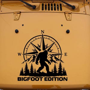 May include: A black vinyl decal applied to a mustard yellow vehicle hood. The design features a compass rose, a silhouette of a mountain range, a forest of pine trees, and a Bigfoot figure. The text BIGFOOT EDITION is printed at the bottom.