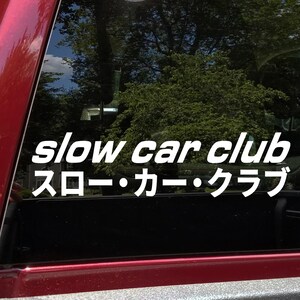 May include: White vinyl decal with the text "slow car club" in a bold, sans-serif font, with Japanese characters below. The decal is affixed to a vehicle window, with a blurred background of trees and sky.