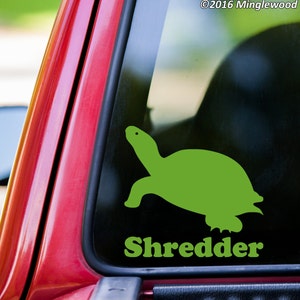 May include: Green turtle silhouette decal with the word "Shredder" underneath. The decal is on a red car window.