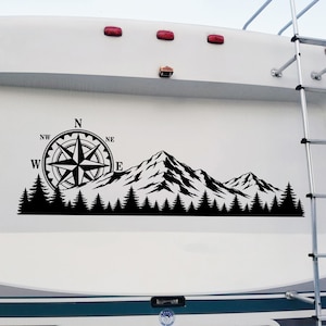 May include: A black vinyl decal applied to the white rear panel of a recreational vehicle. The design features a detailed compass rose with cardinal directions, a silhouette of a mountain range, and a dense forest of pine trees along the base.
