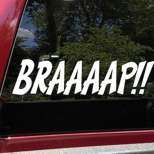 May include: White vinyl decal with the text "BRAAAAP!!" in a bold, comic book style font. The decal is applied to the rear window of a red vehicle.