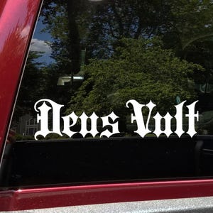 May include: White vinyl decal with the words "Deus Vult" in a gothic font, affixed to a vehicle window. The decal is against a backdrop of trees and a glimpse of a house, with the red frame of the vehicle visible.