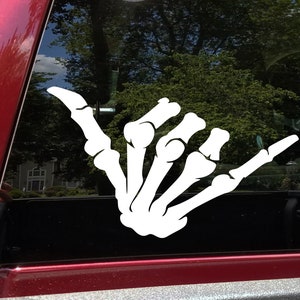 May include: White vinyl decal of a skeleton hand giving the shaka sign. The decal is on a black car window.
