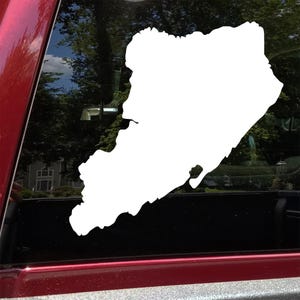 May include: A white vinyl decal in the shape of a geographical outline, possibly a state or county. The decal is affixed to a vehicle window, with a red frame visible. The background shows trees and a house.