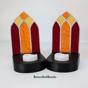 May include: Two stained glass candle holders in a church window design. The holders feature red, orange, and yellow glass panels, with a black base and a white LED candle. The text "BonesAndBevels" is at the bottom.
