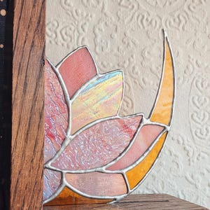 May include: A stained glass bookend featuring a floral design with pink, orange, and yellow hues. The glass is set in a wooden frame. The text "Bones&Bevels" is visible on the base.