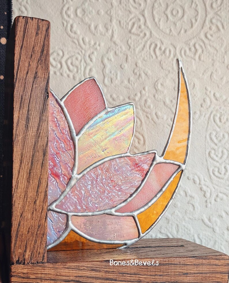 May include: A stained glass bookend featuring a floral design with pink, orange, and yellow hues. The glass is set in a wooden frame. The text "Bones&Bevels" is visible on the base.