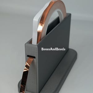 May include: A grey tape dispenser holding rolls of copper foil tape and white tape. The dispenser has the text "BonesAndBevels" on the side. Copper tape is dispensed from the slot.