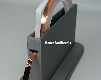 May include: A grey tape dispenser holding rolls of copper foil tape and white tape. The dispenser has the text "BonesAndBevels" on the side. Copper tape is dispensed from the slot.