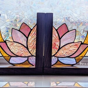 May include: Stained glass lotus flower bookends with a dark wood base and accents. The lotus petals are pink, peach, and gold, with a textured appearance. The bookends are designed to hold books upright. The text "Bones & Bevels" is visible.