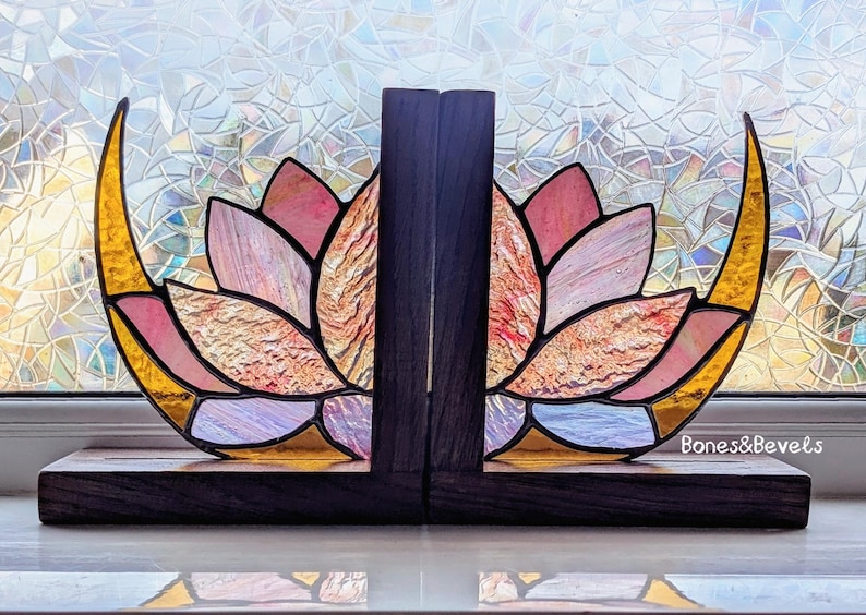 May include: Stained glass lotus flower bookends with a dark wood base and accents. The lotus petals are pink, peach, and gold, with a textured appearance. The bookends are designed to hold books upright. The text "Bones & Bevels" is visible.