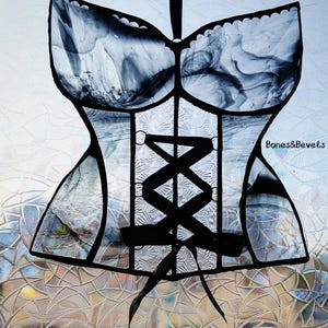 May include: A stained glass corset in shades of gray and black, with a ribbon for hanging. The corset features a detailed design with a lace-up front and the text "Bones&Bevels". The glass has a marbled effect.