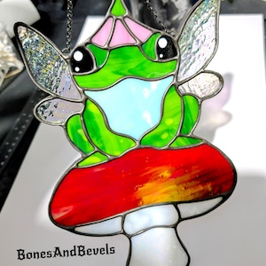 May include: Stained glass frog fairy with iridescent wings, perched on a red mushroom with a white stem. The frog is green with a light blue belly and pink accents. The piece is suspended by a chain and features the text "BonesAndBevels".