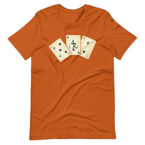 May include: Orange t-shirt featuring three playing cards: the Ace of Spades, the 2 of Spades, and the 9 of Spades. The cards have a vintage, worn look. The shirt is a solid orange color.