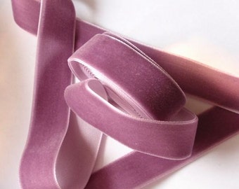 Puede incluir: Primer plano de una cinta de terciopelo malva, parcialmente enrollada y desenrollada. La cinta tiene una textura suave y afelpada y un brillo sutil. Ideal para manualidades y decoración.