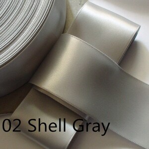 May include: A roll of shell gray satin ribbon. The ribbon is a light, neutral gray color with a smooth, shiny surface. The text "02 Shell Gray" is printed on the ribbon.