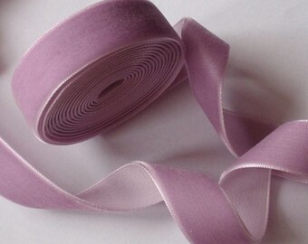 Puede incluir: Un rollo de cinta de terciopelo lavanda con reverso de satén. La cinta está desenrollada, revelando su textura y color. Ideal para manualidades, costura o envolver regalos.