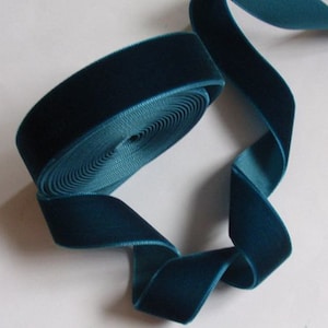 Puede incluir: Un rollo de cinta de terciopelo color teal con una textura suave y afelpada. La cinta está desenrollada, revelando un reverso liso similar al satén. El color de la cinta es un teal profundo y rico, perfecto para proyectos de manualidades y decoración.