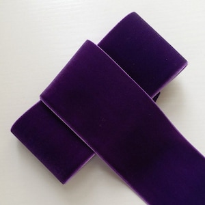 May include: A length of deep purple velvet ribbon, folded over itself. The image highlights the soft texture and rich colour of the fabric. The ribbon has a slight sheen, and the edges are neatly finished.