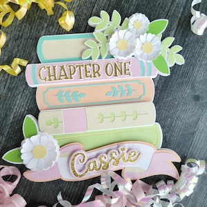 May include: A layered paper craft decoration featuring a stack of books in pastel shades of teal, peach, pink, and green. The top book spine reads CHAPTER ONE in gold glitter, and a pink banner at the bottom displays the name Cassie in gold.