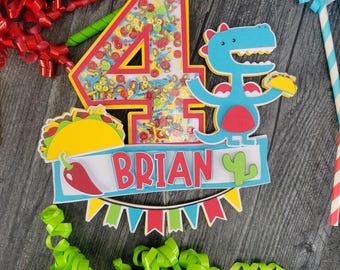 May include: A colorful birthday cake topper featuring the number 4 with confetti, a blue dinosaur holding a taco, and a banner that says "BRIAN". The topper includes a taco and a cactus.