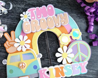 May include: A colorful cake topper with a retro theme. It features a rainbow, a peace sign, a flower, and a van. The text reads "Two Groovy" and "Kinsey". The topper is decorated with daisies and sunglasses.