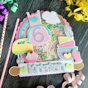 May include: A multi-layered cake topper with a pink and teal color scheme. The design includes a dress with the number 6, a chef's hat, a whisk, bowls, a mixer, a cupcake, and the name "Kristen". Sprinkles are visible.