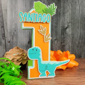 May include: A colorful birthday cake topper in the shape of the number one, with the name "SANTIAGO" in teal letters. The topper features a teal dinosaur, dinosaur footprints, and tropical leaves. The background is orange and green.