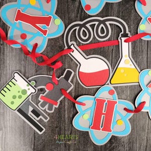 May include: A science-themed banner with cut-out shapes of atoms, beakers, and a microscope. The banner features the letters "Y" and "H" in red, with a red ribbon strung through the cutouts. The color scheme includes gray, blue, red, yellow, and green.