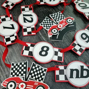 May include: A red, black, and white race car themed banner with numbers 4-12, checkered flags, and race cars. The banner is strung on a red ribbon, perfect for a birthday party or race car themed event.