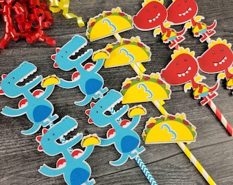 May include: A collection of colorful party decorations, including dinosaur and taco-themed cupcake toppers. The toppers feature blue dinosaurs, yellow tacos with the number 3, and red dinosaur heads. Ribbons in yellow, red, and green are scattered around.