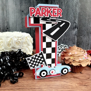 May include: A red, white, and blue number one cake topper with the name "PARKER" on top. The topper has a checkered flag, a race car, and a road design. A cupcake with chocolate frosting is next to the topper.