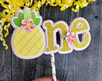 May include: A layered cake topper with a pineapple and the word "one". The pineapple is yellow with a textured design, green leaves, and a pink and yellow flower. The word "one" is gold glitter with a pink outline and a small flower. The stick has a flamingo pattern.