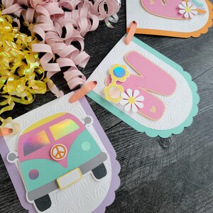 May include: A close-up of three decorative paper banners with a retro theme. One banner features a colorful van with a peace sign, while the others display the letter "N" and a daisy. The banners are in pastel colors with scalloped edges.